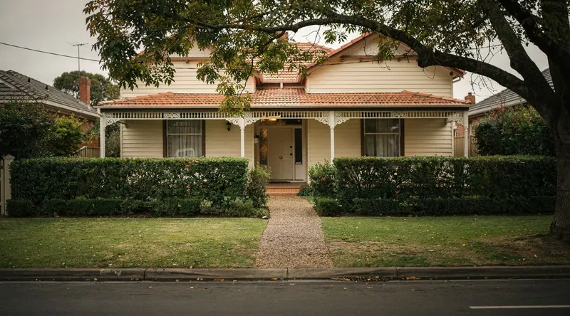 A weatherboard home with a clipped front hedge and mown lawn on a leafy street